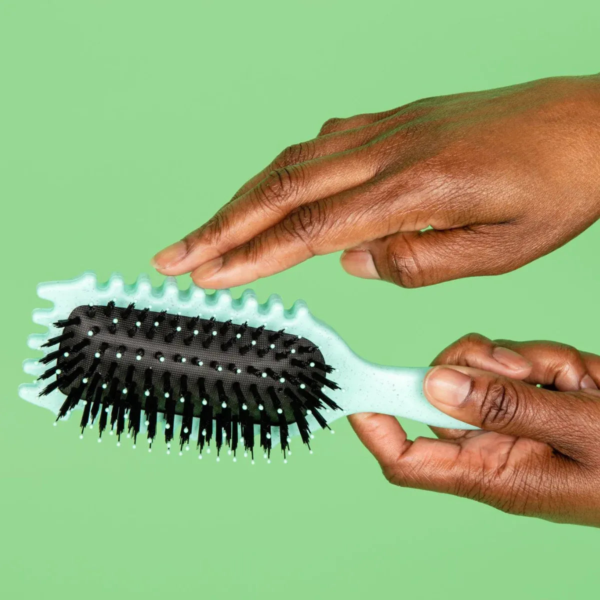 Mint green CurlPro Define Styling Brush with lateral grooves for curl definition, held by hands against a green backdrop.