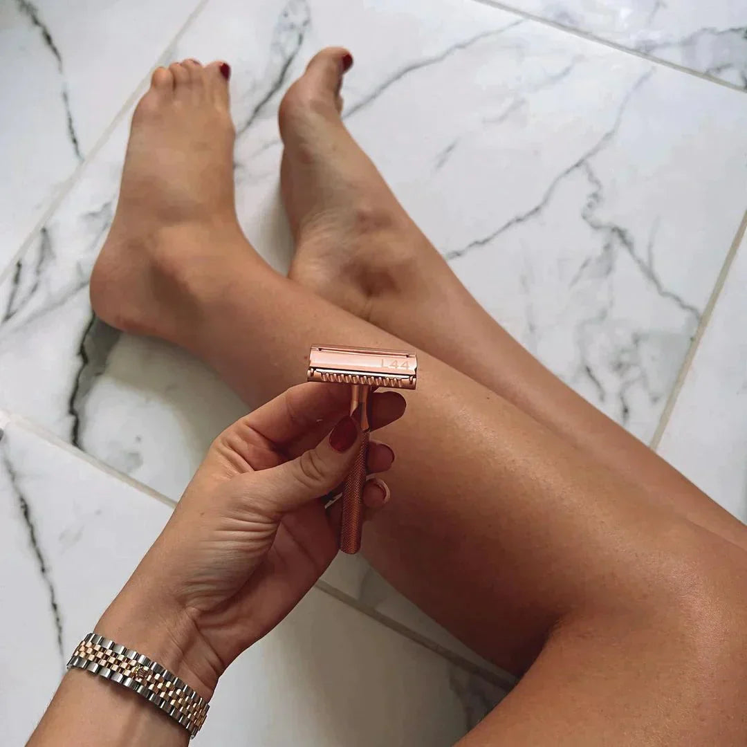 Rose gold SmoothShave safety razor held against legs for a smooth shave on a white marble background.
